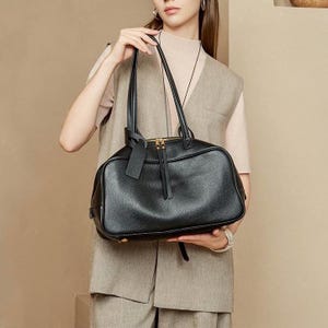 May include: Black leather handbag with a rectangular shape and dual handles. The bag features a top zipper closure with gold-toned hardware and a small, attached tag. The bag is held by a person wearing a neutral-toned outfit.