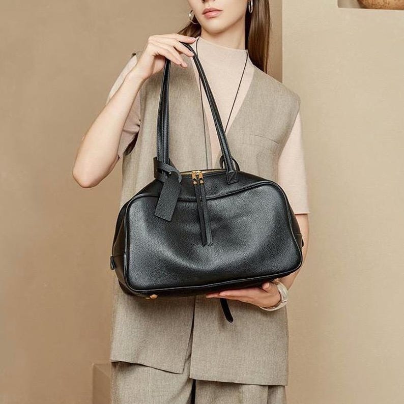 May include: Black leather handbag with a rectangular shape and dual handles. The bag features a top zipper closure with gold-toned hardware and a small, attached tag. The bag is held by a person wearing a neutral-toned outfit.