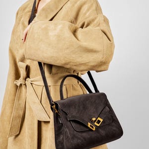 May include: A dark brown leather handbag with a top handle and a long shoulder strap. The bag features a flap closure with gold-colored hardware. The person is wearing a tan suede coat.