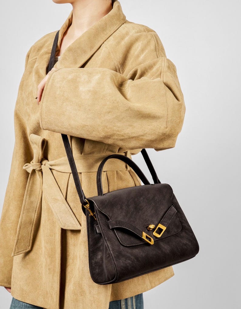 May include: A dark brown leather handbag with a top handle and a long shoulder strap. The bag features a flap closure with gold-colored hardware. The person is wearing a tan suede coat.