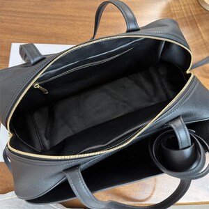 May include: Open view of a black leather handbag with gold-colored zippers and handles. The bag has a rectangular shape with a spacious interior, featuring a smaller zippered pocket. The handles are sturdy and the bag appears to be new.