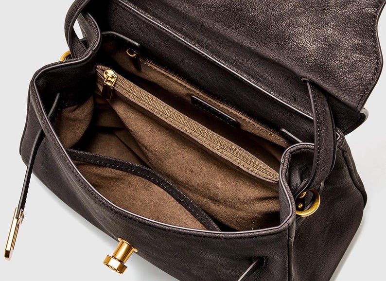 May include: Open view of a dark brown leather handbag with a flap closure. The interior features a zippered pocket and multiple compartments. Gold-toned hardware accents the bag, including the clasp and handle attachments.