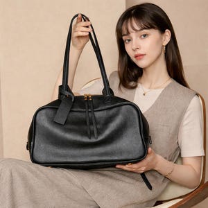 May include: A black leather handbag with a textured surface, featuring two long shoulder straps, a top zipper closure with gold-toned hardware, and a matching rectangular leather luggage tag attached to one of the handles.