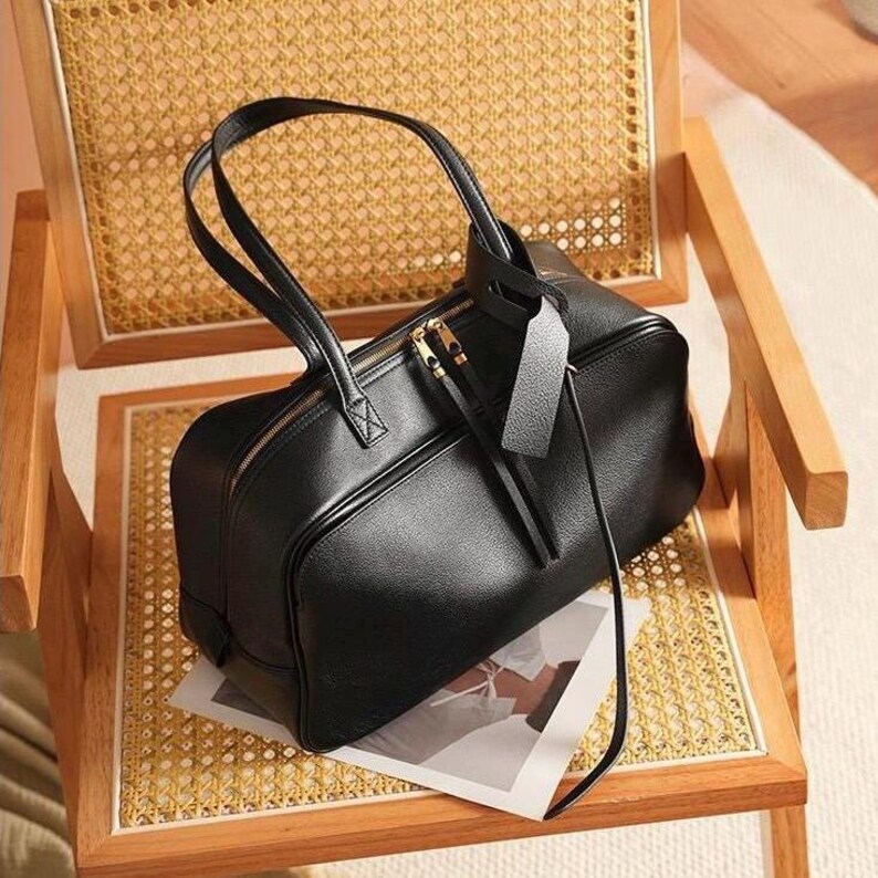 May include: A black leather handbag with a textured surface, featuring two top handles, a gold-toned zipper closure, and a rectangular leather luggage tag attached to one handle. The bag has a structured, rectangular shape with rounded corners.