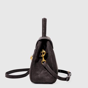 May include: A dark brown leather handbag with a top handle and a detachable shoulder strap. The bag features gold-colored hardware, including a clasp closure and strap attachments. The bag is designed with a structured shape.