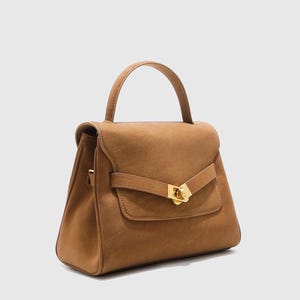 May include: A tan handbag with a top handle and a flap closure. The bag features a gold-colored clasp and a small front pocket. The bag is made of a suede-like material and has a structured shape.