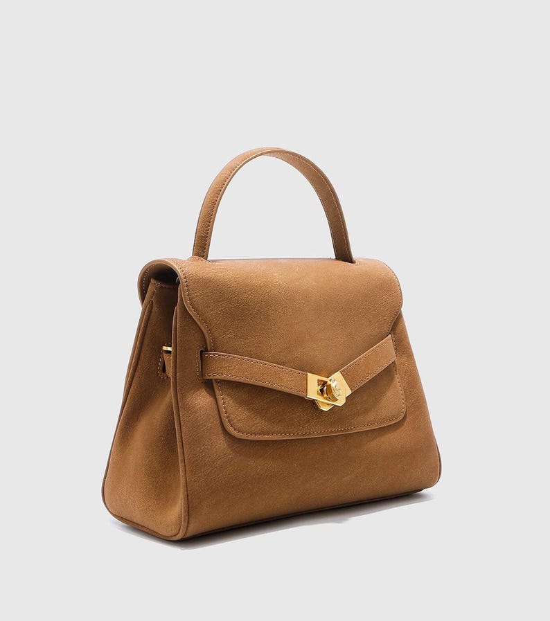 May include: A tan handbag with a top handle and a flap closure. The bag features a gold-colored clasp and a small front pocket. The bag is made of a suede-like material and has a structured shape.