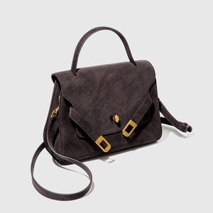 May include: A dark gray handbag with a top handle and a long, adjustable shoulder strap. The bag features a flap closure with a gold-colored clasp and two gold-colored rectangular buckles. The bag's material appears to be suede or a similar textured fabric.