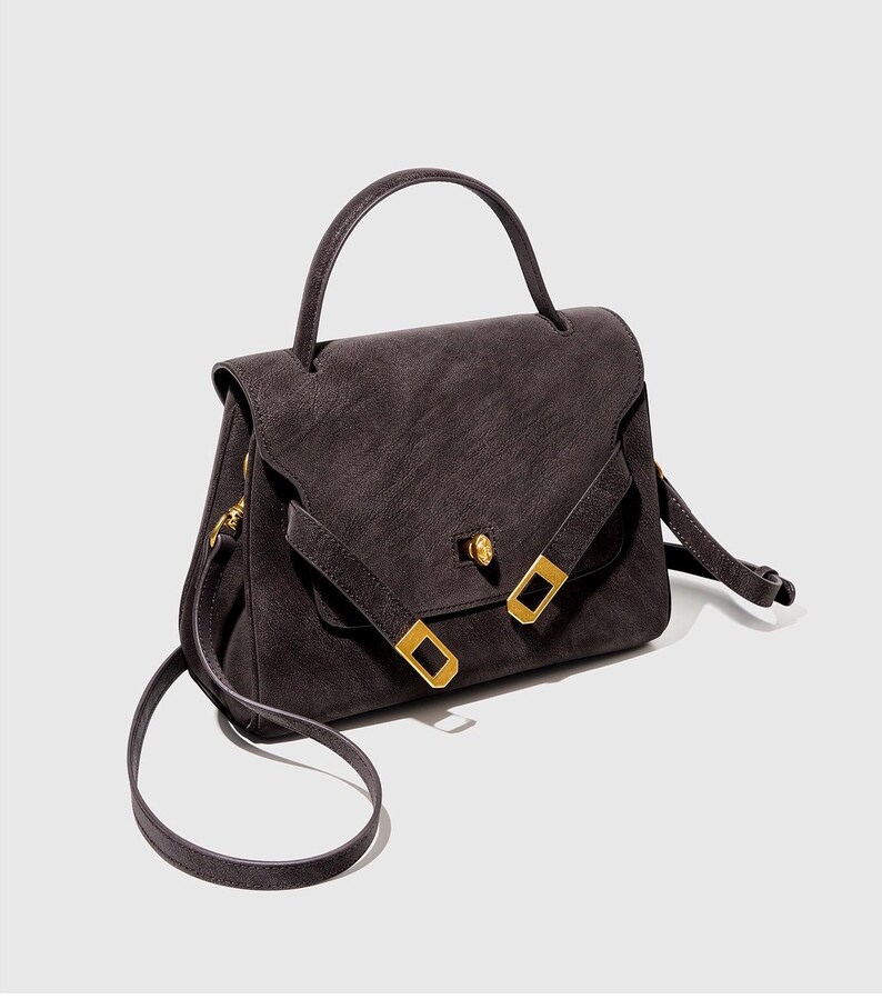 May include: A dark gray handbag with a top handle and a long, adjustable shoulder strap. The bag features a flap closure with a gold-colored clasp and two gold-colored rectangular buckles. The bag's material appears to be suede or a similar textured fabric.