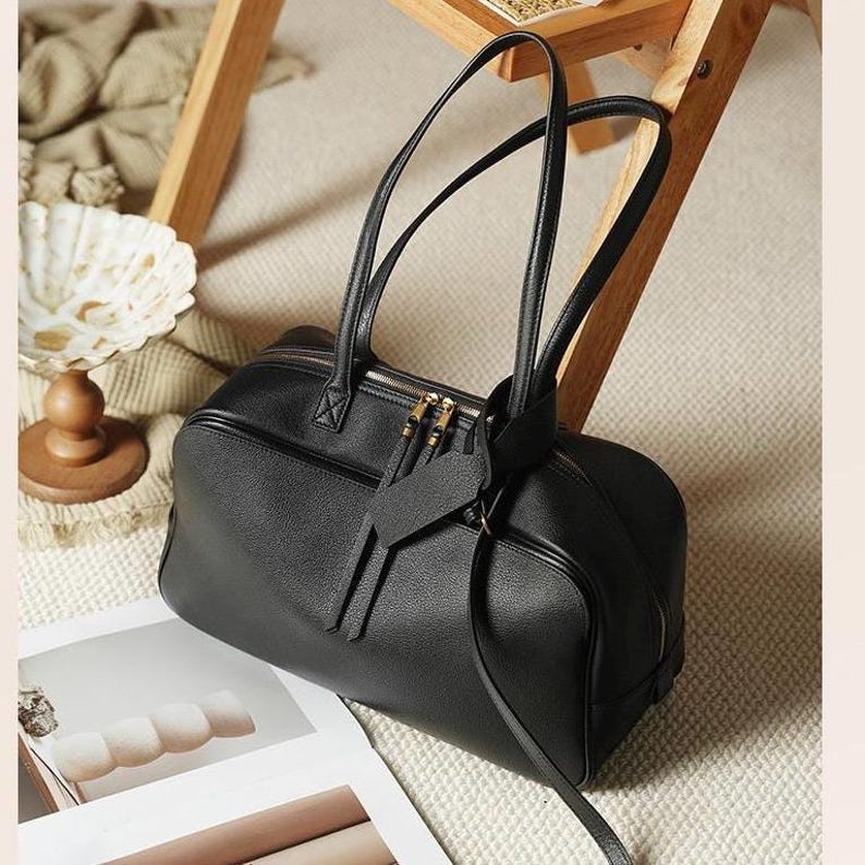 May include: A black leather duffel bag with two long handles and a zipper closure. The bag has a rectangular shape with rounded edges and a small tag attached to the zipper. The bag is sitting on a light-colored surface.
