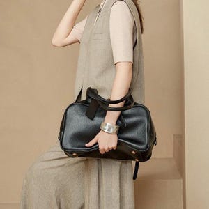 May include: A black leather handbag with a rectangular shape, featuring a top handle and a detachable shoulder strap. The bag has gold-colored studs on the bottom and a small tag attached to the handle. The bag is held by a person wearing a silver bracelet.