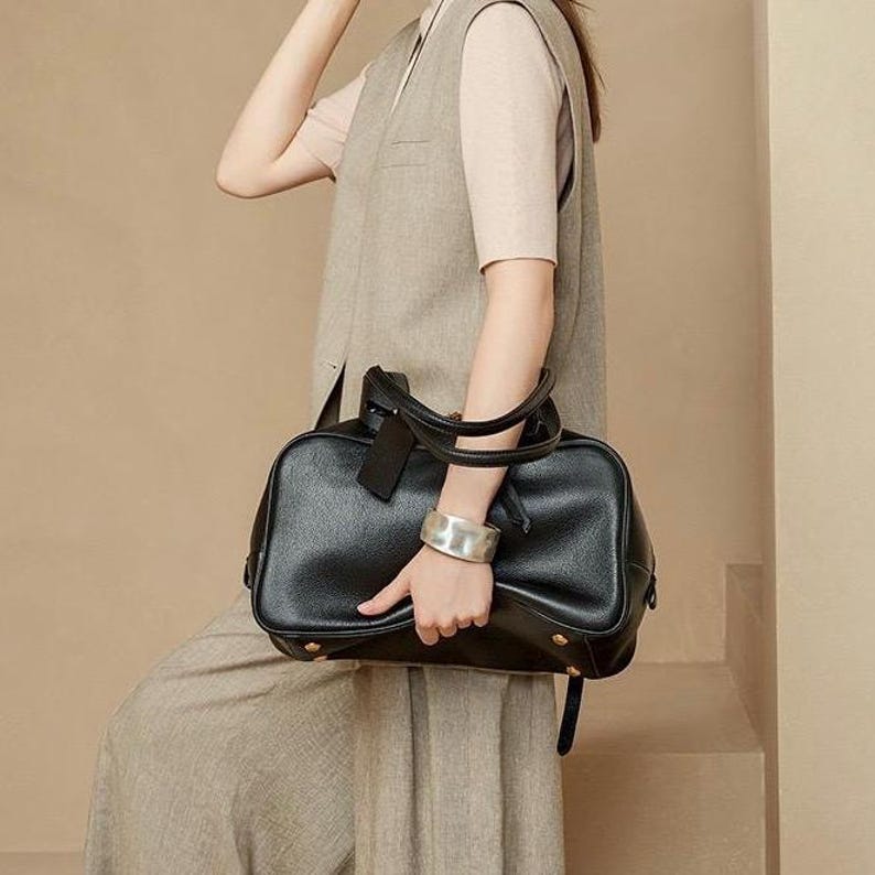 May include: A black leather handbag with a rectangular shape, featuring a top handle and a detachable shoulder strap. The bag has gold-colored studs on the bottom and a small tag attached to the handle. The bag is held by a person wearing a silver bracelet.