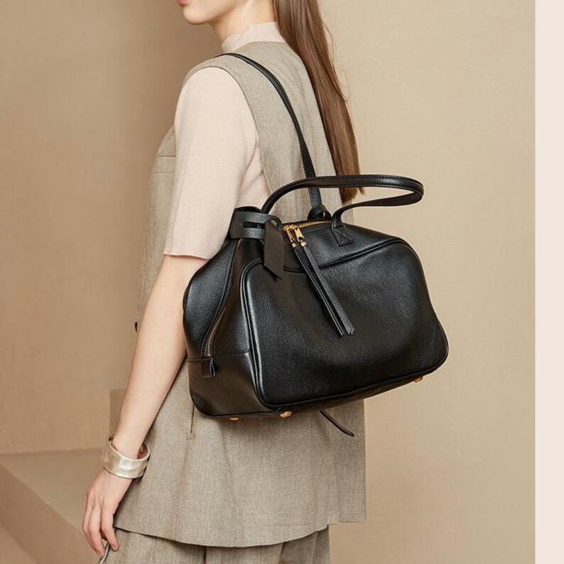 May include: A black leather handbag with a top handle and shoulder straps. The bag has a zippered closure and a tassel detail. The bag is worn over the shoulder. The bag is a dark color.