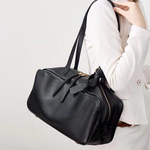 May include: Black leather satchel bag with a rectangular shape. The bag has a top handle and a shoulder strap. It features a gold-colored zipper and a decorative leather tie. The bag is carried over the shoulder.