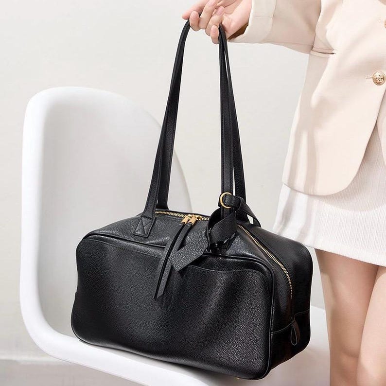 May include: A black rectangular handbag with a textured surface, featuring two long shoulder straps, a gold-toned top zipper closure with dual pulls, and a matching black leather-like luggage tag attached to a gold-toned side ring.