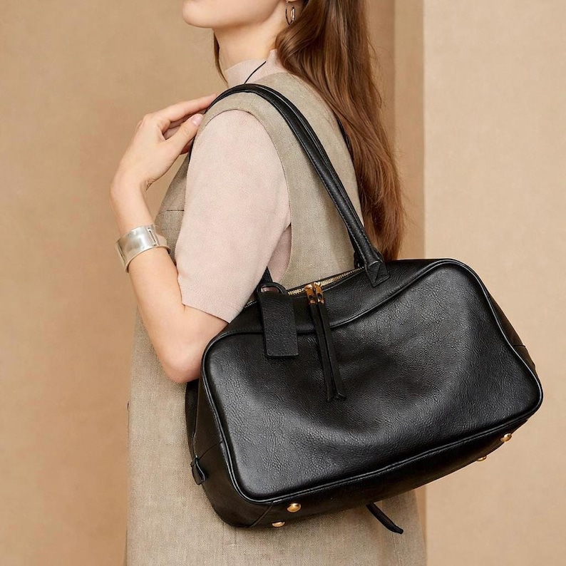 May include: Black leather handbag with a rectangular shape, featuring a top zipper closure and dual handles. The bag has gold-colored metal accents on the base and zipper. A small leather tag hangs from the zipper.