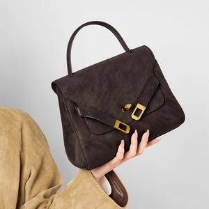 May include: A dark brown handbag with a top handle and a flap closure. The bag features gold-colored hardware on the flap and a structured design. The bag is held by a person wearing a tan jacket.