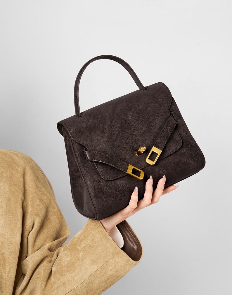 May include: A dark brown handbag with a top handle and a flap closure. The bag features gold-colored hardware on the flap and a structured design. The bag is held by a person wearing a tan jacket.