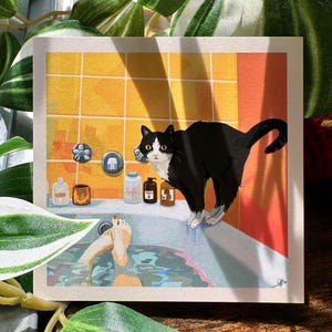 May include: A square art print featuring a black and white cat standing on the edge of a bathtub. Inside the tub, a pair of feet are visible in blue water. The background shows yellow tiled walls with various bottles and a faucet fixture.