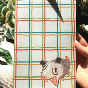 Peut inclure: Un bloc-notes rectangulaire avec un fond blanc et un motif à carreaux coloré rouge, vert, bleu et orange. Une illustration de dessin animé d'un opossum est dans le coin inférieur droit. Le bloc-notes est tenu par une main.