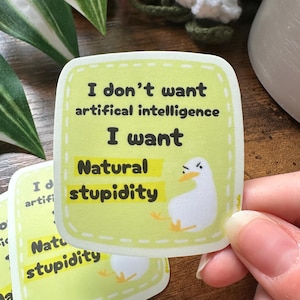 Puede incluir: Una pegatina verde claro con el texto "No quiero inteligencia artificial, quiero estupidez natural". Se muestra un pato de dibujos animados. La pegatina es cuadrada con esquinas redondeadas y un borde punteado.