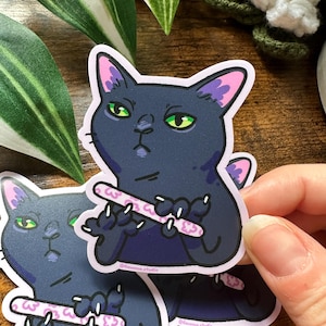 May include: A sticker featuring a cartoon black cat with green eyes, holding a pink and white nail file. The cat has purple accents on its ears and is set against a white background. The sticker has the text "@devenue.studio".
