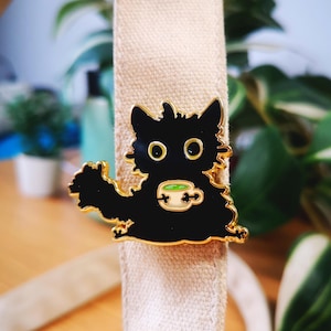 Peut inclure: Épingle en émail noire en forme de chat, bordée d'or, tenant une tasse de thé avec un liquide vert. L'épingle est posée sur un fond beige avec des plantes vertes. Le chat a de grands yeux ronds et jaunes.