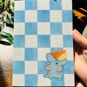 Könnte beinhalten: Ein Notizbuch mit einem blau-weißen Schachbrettmuster. Auf dem Cover befindet sich eine Cartoon-Maus mit einem Käse auf dem Kopf. Das Notizbuch wird von einer Hand gehalten.