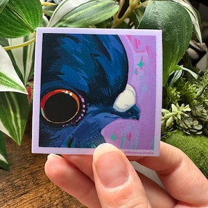May include: A square sticker featuring a close-up illustration of a blue creature with a large, circular eye. The eye is black with a gold and red outline. The background is purple with pink and green accents. The sticker has a white border and the text "@6avenue.studio".