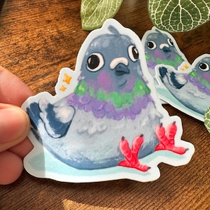May include: A sticker featuring a cartoon pigeon with a blue, purple, and green design. The pigeon has large black eyes, red feet, and is surrounded by small yellow stars. Additional pigeon stickers are visible in the background.