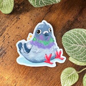 May include: A sticker featuring a cartoon pigeon with a blue head, purple and green chest, and red feet. The pigeon is sitting on a light blue base. The sticker has a white border and is surrounded by green leaves.