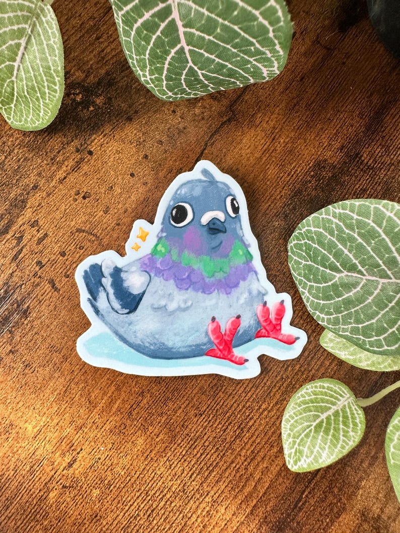 May include: A sticker featuring a cartoon pigeon with a blue head, purple and green chest, and red feet. The pigeon is sitting on a light blue base. The sticker has a white border and is surrounded by green leaves.