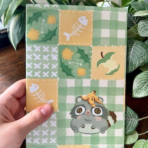 Può includere: Un taccuino rettangolare con un design stravagante. La copertina presenta un motivo patchwork con quadrati gialli, quadretti verdi e illustrazioni di lische di pesce, fiori e un procione con una buccia di banana sulla testa.