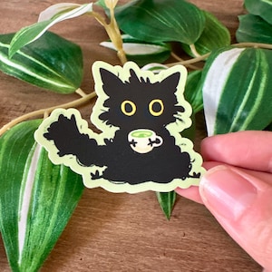 May include: A die-cut sticker featuring a fluffy black cat with large yellow eyes holding a small white teacup filled with a green beverage. The cat has a shaggy, unkempt appearance and is outlined in a pale yellow border against a light background.