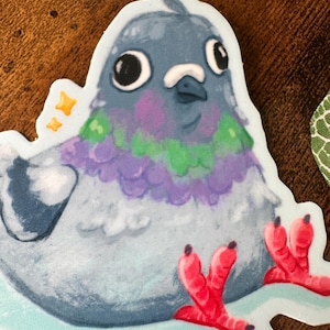 May include: A sticker featuring a cartoon pigeon with a blue-grey body, purple and green neck feathers, and bright red feet. The pigeon has large, expressive eyes and is surrounded by a white border with two small yellow stars.