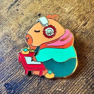 May include: A colorful enamel pin featuring a cartoon capybara wearing headphones and a hoodie, seated at a red desk. The pin has gold-colored trim and the word "lofi" is visible on the headphones.