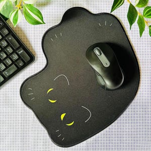 Peut inclure: Tapis de souris noir en forme de chat avec des yeux jaunes et des moustaches blanches. Une souris sans fil noire est posée dessus. Le tapis est sur une surface à carreaux, avec un clavier et de la verdure en arrière-plan.