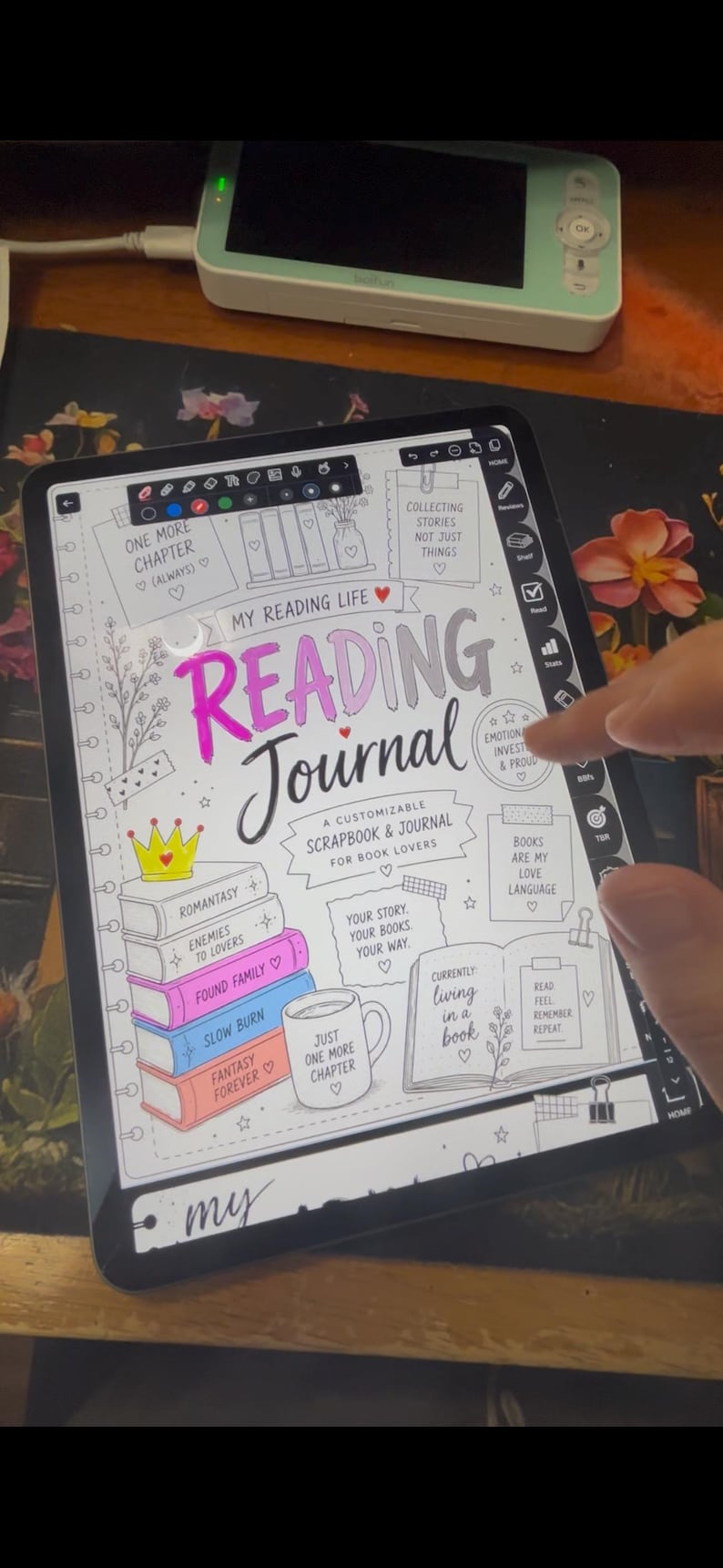 May include: A digital reading journal on a tablet, with the title "Reading Journal" in pink and black. The journal includes prompts, book stacks, and the text "A Customizable Scrapbook & Journal for Book Lovers."