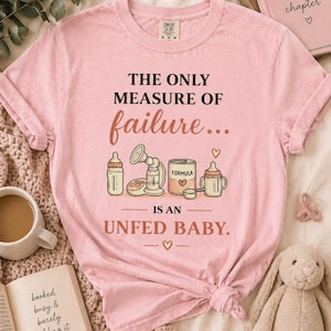 May include: A pale pink t-shirt with the words "THE ONLY MEASURE OF failure... IS AN UNFED BABY." The design includes illustrations of baby bottles, a breast pump, and formula. A book, a cup of coffee, and a toy bunny are also present.