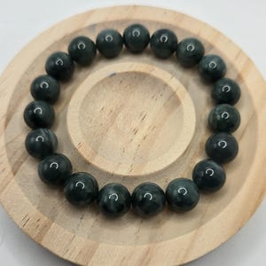 May include: A dark green beaded bracelet with a polished finish. The round beads are strung together, forming a complete circle. The bracelet is displayed on a small, round wooden tray.