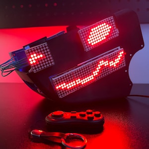 May include: A black mask featuring three red LED dot matrix displays showing illuminated patterns, connected by blue wires. Below the mask sits a small black handheld game controller and a metal keychain with a black leather strap.