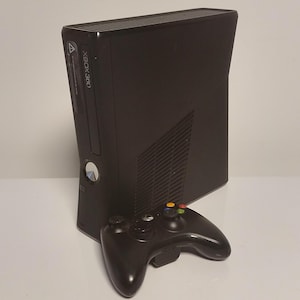 Könnte beinhalten: Schwarze Xbox 360 Konsole mit passendem schwarzem Controller. Die Konsole steht aufrecht, mit dem Xbox 360 Logo sichtbar. Der Controller hat farbige Knöpfe und zwei Analogsticks.