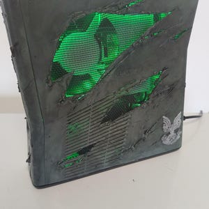 Könnte beinhalten: Eine modifizierte Xbox 360-Konsole mit einem abgenutzten, kampferprobten Aussehen. Das graue Gehäuse weist zerrissene Abschnitte auf, die einen grün beleuchteten Innenraum freilegen. Ein silbernes Emblem ist auf der Seite sichtbar. Die Konsole ist angeschlossen.