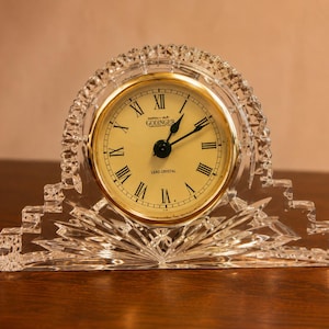 May include: A clear crystal desk clock with a gold-tone face and black hands. The clock features Roman numerals and the brand name "Godinger". The clock is set in a decorative, faceted crystal frame.