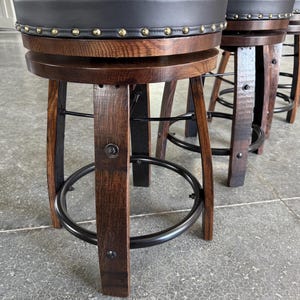 May include: A set of swivel bar stools featuring dark brown curved wooden legs, a circular black metal footrest, and a round seat upholstered in black material with a decorative border of brass-coloured nailhead trim along the edge.
