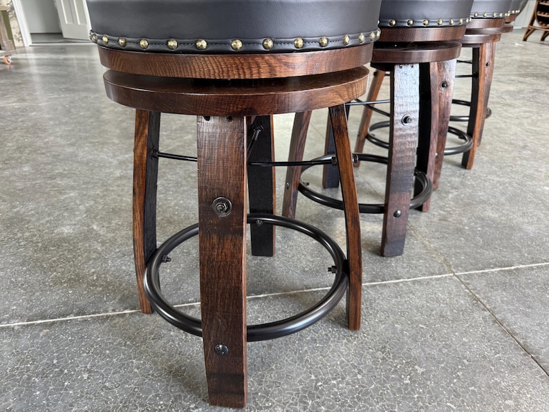 May include: A set of swivel bar stools featuring dark brown curved wooden legs, a circular black metal footrest, and a round seat upholstered in black material with a decorative border of brass-coloured nailhead trim along the edge.
