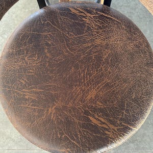 May include: A circular stool seat featuring a dark brown surface with a distressed, textured finish that reveals lighter tan undertones. The material displays a cracked, weathered pattern across the entire round top, supported by a black metal frame.