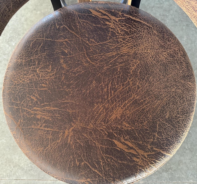 May include: A circular stool seat featuring a dark brown surface with a distressed, textured finish that reveals lighter tan undertones. The material displays a cracked, weathered pattern across the entire round top, supported by a black metal frame.