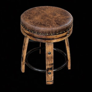 May include: A round stool featuring a distressed brown textured seat with a row of brass-coloured nailhead trim. The base is constructed from light-toned wood legs with a dark metal circular footrest and metal hardware accents on each leg.