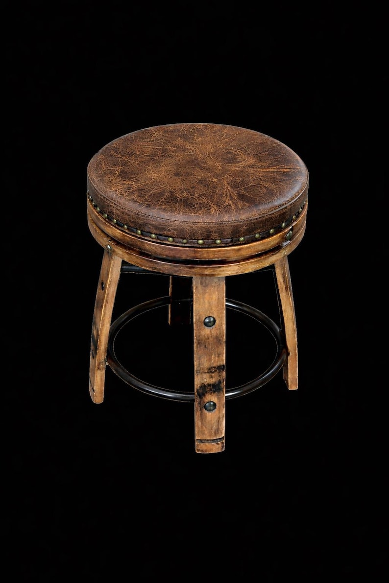 May include: A round stool featuring a distressed brown textured seat with a row of brass-coloured nailhead trim. The base is constructed from light-toned wood legs with a dark metal circular footrest and metal hardware accents on each leg.